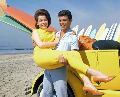 Frankie Avalon holding Annette Funicello on beach next to dune buggy with surfboards, sand ocean in background