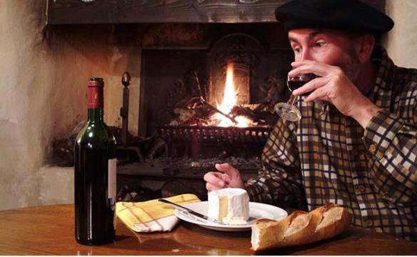 man with a beret drinking wine at a table with bottle of wine glass of wine cheese and baguette in front of open fire, Frankreich, France, Franzose, french