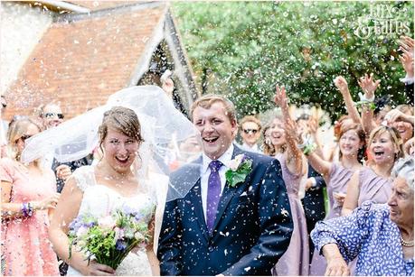 bride and groom smile at wedding whilst confetti is thrown at them bride is holding a wildflower bouquet 