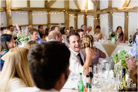 Guest at UK barn wedding smiles and laughs during the speeches with fairy lights strung up behing him 