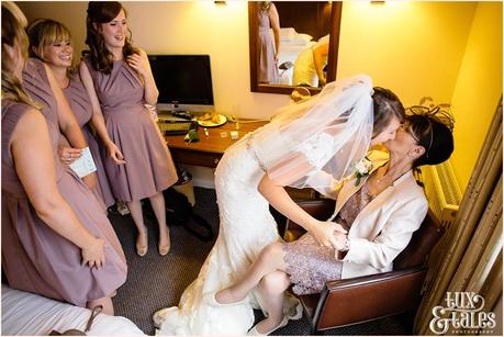 Bride kisses mother who is sitting in a chair by the window