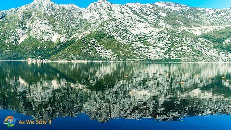 Mountains reflected in the smooth water of the bay. More fish farm floats in the foreground.