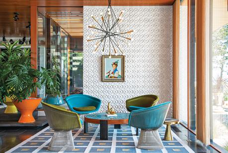 white ceramic tiles cover the back wall while green and blue chair sit around a coffee table with a blue mosaic surface on a rug with a pattern of blue squares 
