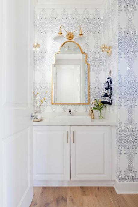 Elegant printed wallpaper in blue and white with gold accents and mirror above the sink with white bathroom cabinets. Stunning bathroom design idea!: 