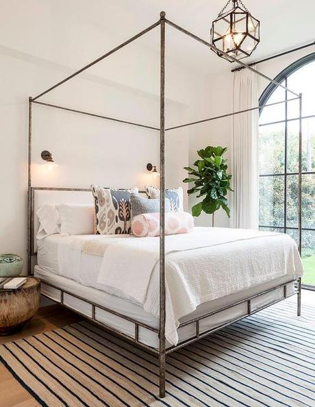 Chic bedroom features a metal canopy bed, Oly Studio Marco Bed, dressed in soft white bedding and pink and gray ikat pillows as well as a pink ikat bolster pillow placed atop a beige and navy striped rug illuminated by glass vintage barn wall sconces and a Suzanne Kasler Morris Lantern.: 