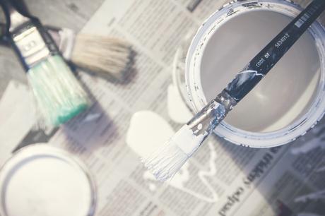 A tin of paint with a paintbrush on top sitting on a pile of newspapers with some more paintbrushes on the ground ready to use