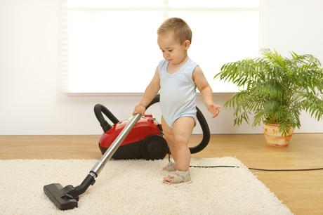Small baby vacuuming a rug in a sitting room with a plant in the background