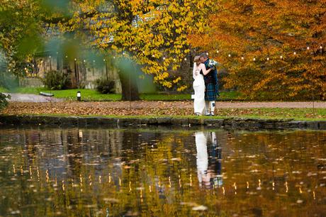 Bride and groom wearing kilt and tartan kiss in autumn colours by lake at East Riddlesden Hall Wedding