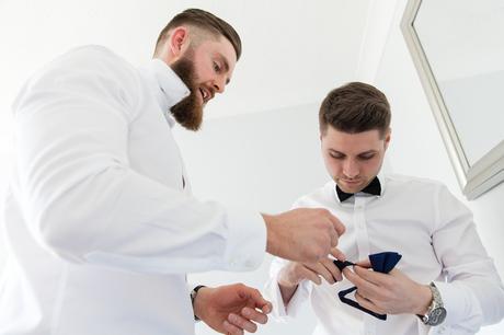 Best man and groom with beard try to figure out how to tie their bow tie. 