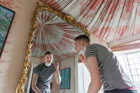 Groom shaving whilst looking into a mirror. French fabric hangs fromt he ceiling. 