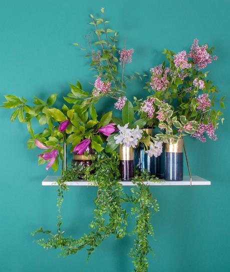 There are five glass vases on this shelf of varying heights and colour and they are filled, very simply, with whatever is growing in the garden. Branches of magnolia, weigela and lilac for height with flower heads of rhododendrons for the smaller vases. An artificial trailing fern completes the look and extends the vignette for more interest.