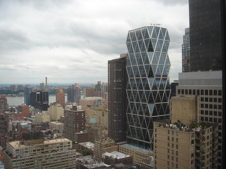 76. The Hearst Tower, a sleek modern skyscraper inspired by the 1920s on New York City's Eighth Avenue, stands majestic amid drab rectangular buildings.