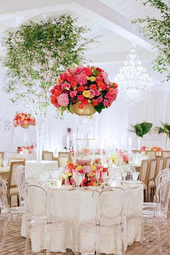 tall wedding centerpieces on a round table with a white tablecloth surrounded by flowers on a transparent stand in a hollow vase bright red flowers carrie patterson via instagram