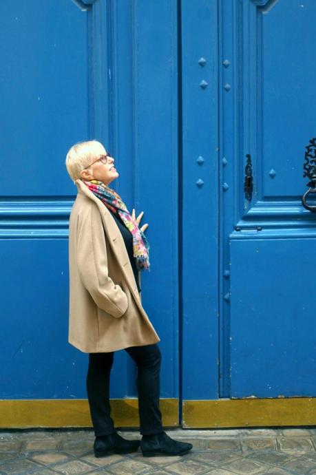 Susan B. wears a camel jacket from agnes b. in front of a blue door in Paris.
