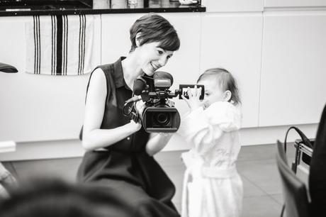 Female wedding videographer is crouched down with a smile to show a little flower girl how her Mummy the bride looks on her video camera as they get ready for a wedding near Manchester