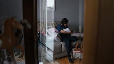 A mom sits on her gray leather sofa breastfeeding her newborn daughter