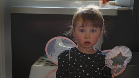 a pretty little 3 year old girl kneels on the floor looking past the family photographer wearing her fairy wings and hold a pink wand with bright red rosy cheeks