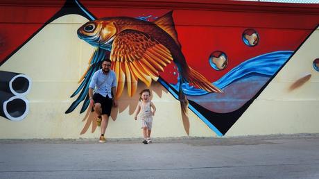 A little girl runs excitedly towards to camera as her Dad stands by a dramatic flying fish graffiti art on the streets of Lisbon