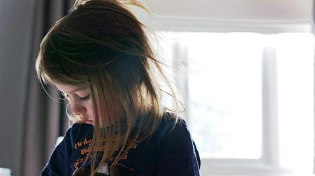 a photograph of a young girl sat in front of a bright window looking down with her hair all messy from a good nights sleep