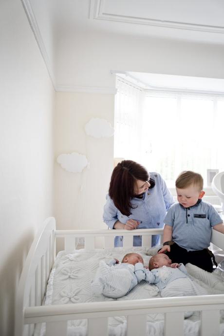 a mom leans over to chat to her toddler about their new baby boy twins in their bright and neutral white and cream nursery in the wirral