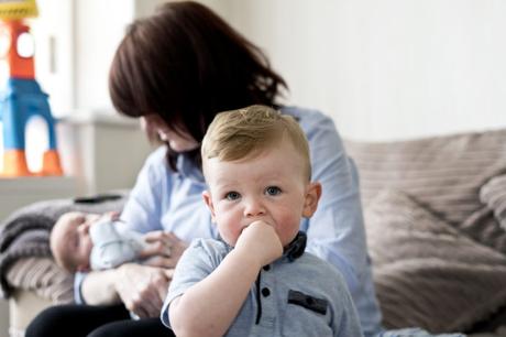 a little boy chews his hand as he looks at the camera His mom is holding her newborn son on the sofa behind him