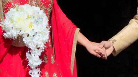 the bangladeshi wedding saree in red looks vibrant in the bright sunshine for the wedding video. The bride and groom are holding hands