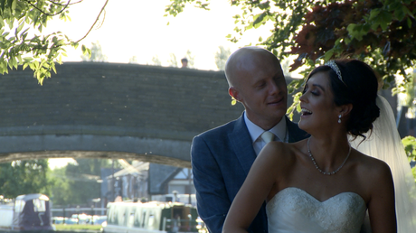 the groom smiles at his laughing bride as they pose for the wedding video and photos outside the blue mallard alongside the leeds liverpool canal in Burscough