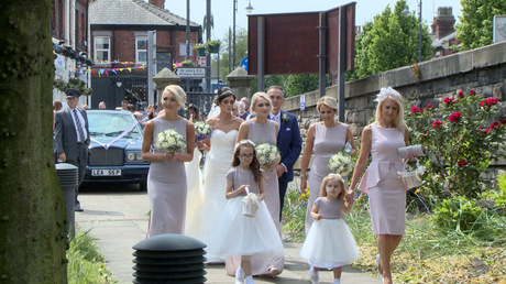 the bride and her bridal party walk down the stone path to make their way in to st johns church in Burscough for the wedding ceremony the bridesmaids are wearing full length lilac purple dresses and holding cream flowes
