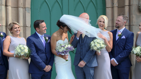 the brides veil gets blown in front of the grooms face during their wedding photos outside st johns church in burscough