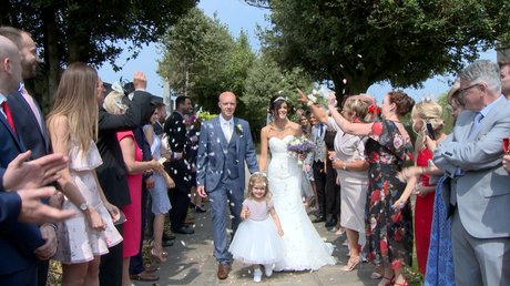 a flower girl walks and laughs in front of the bride and groom as they walk towards the wedding photogapher and videographer for their confetti photo