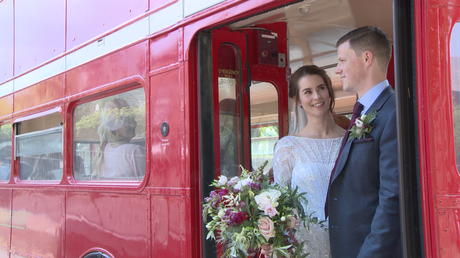 the bride and groom chat standing on the back of the red london bus after their wedding ceremony at St Peters Church