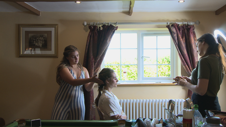 a wide shot shows the bride sitting in front of her living room window as the bridal hair and make up team add the finishing touches before she gets ready for her wedding in Liverpool