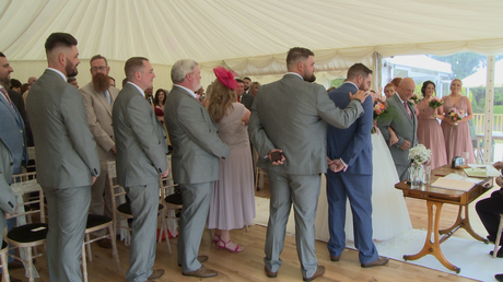 the best man reassuringly pats the groom on the shoulder as his bride arrives at the top of the aisle at the lakeside marquee