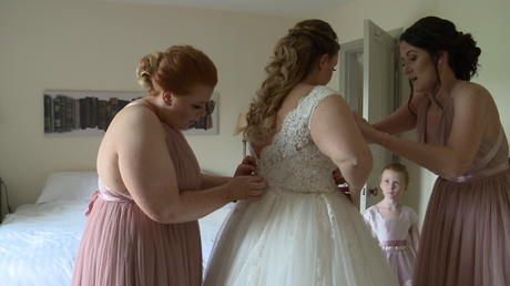 two bridesmaids help the bride fit in to her wedding dress as a flower girl looks on