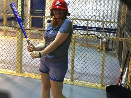 Rach in a batting cage with baseball bat in hand and helmet on ready to play!
