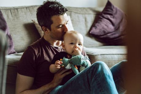 dad sits on the floor next to their grey sofa at home in Sale and sneaks a kiss on top of his sons forehead 