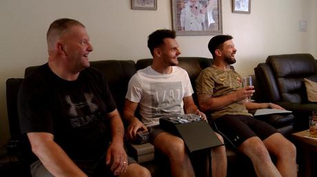 the groom sits on a sofa between his Dad and best man as he opens gifts from his bride for the wedding video in wirral