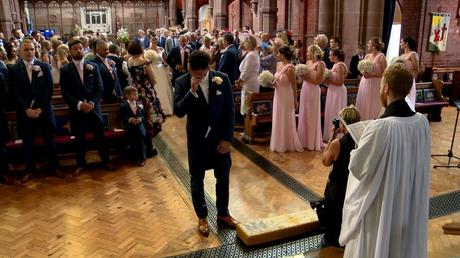 the groom wipes a tear away with his hand after seeing his bride walk down the aisle in her classic Ellis Bridal dress with her dad all caught on video by love gets sweeter