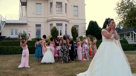 the bride covers her face as she laughs seeing her friends fight for her wedding bouquet in front of the wedding videographer outside thornton Halls white main building