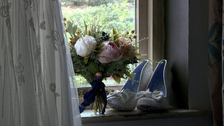 a pair of white glitter irregular choice bridal shoes sit on a window sill beside the brides dress and artificial peony bouquet