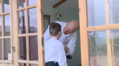 A dad helps fasten the buttons of his sons shirt before his wedding at abels harp the videographer is standing outside the pod as they get dressed inside
