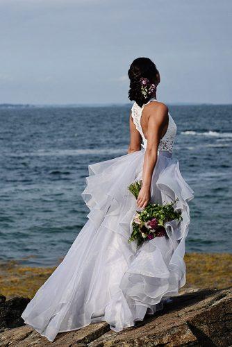 beach styled shoot bride with wedding bouquet flower updo in a lush white dress on the rocks against the sea alexi