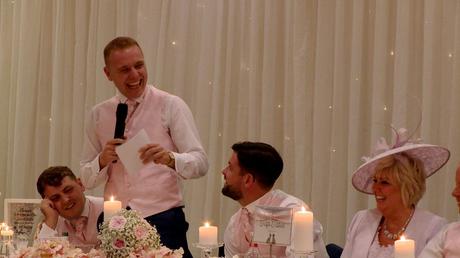 a best man laughs during his speech in front of a sparkly lit white backdrop during a wedding at Thornton Hall