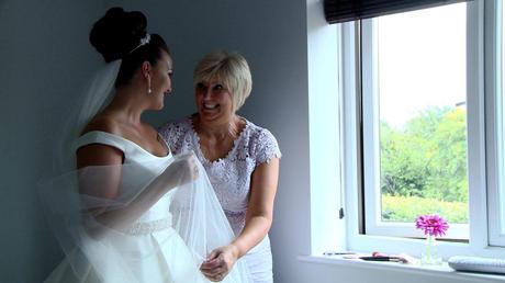 the mother of the bride shrugs her shoulds in excitement as she helps her daughter with the finishing touches to her wedding outfit with the videographer and photographer capturing the moment in their home in the wirral