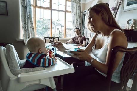dad looks on from the corner of their living room in Sale as Mum spoon feeds their little boy his dinner whilst the family photographer snaps away