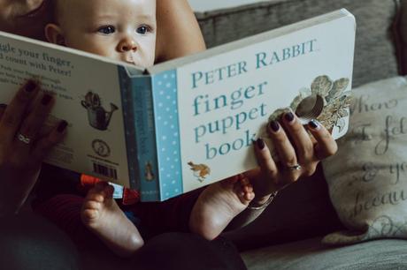 a little boy sits on his Mum's knee as they read a peter rabbit finger puppet book. You can see his eyes peering over the top and his little feet under the book. from a family photo shoot in Sale Manchester