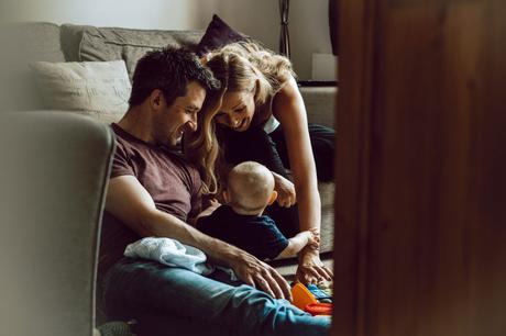 a dad sits on the floor of their living room in Sale Manchester next to his 7 month old little boy as Mum leans in with a huge smile to make him smile as the photographer sits by the door capturing a relaxed family moment at home