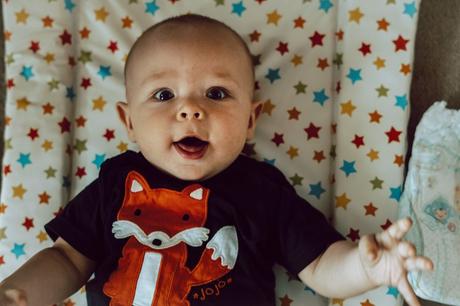 A blue eyed baby boy is wide eyed staring up at the family photographer with a navy fox t shirt on laying on his colourful star changing mat at home