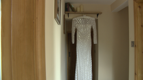 A long floor length beaded Eliza Jane Howell Gown hangs from a small bookshelf by a door on a cream silk bridal hanger
