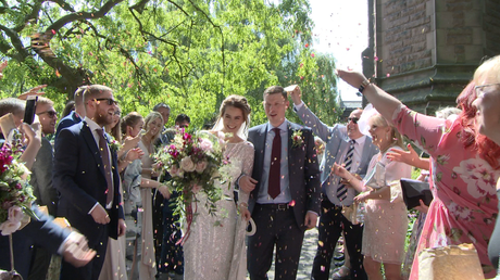an Eliza Jane Howell bride holds a pink and purple rustic hand tied oversized bouquet holding her dress and linking her grooms arm as they get showered in confetti outside St Peter's Church for their Liverpool wedding